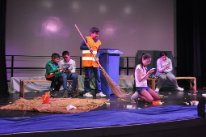 Ein Junge trägt eine Leuchtweste und wischt mit einem grossen Besen, Abfall zusammen. Im Hintergrund sitzen weitere Kinder.